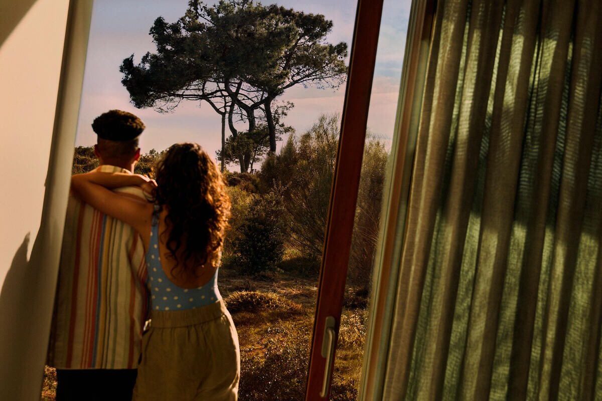 A young pair in colorful, patterned clothing faces away from the camera looking out a sliding door at the desert surrounding the vacation rental property.